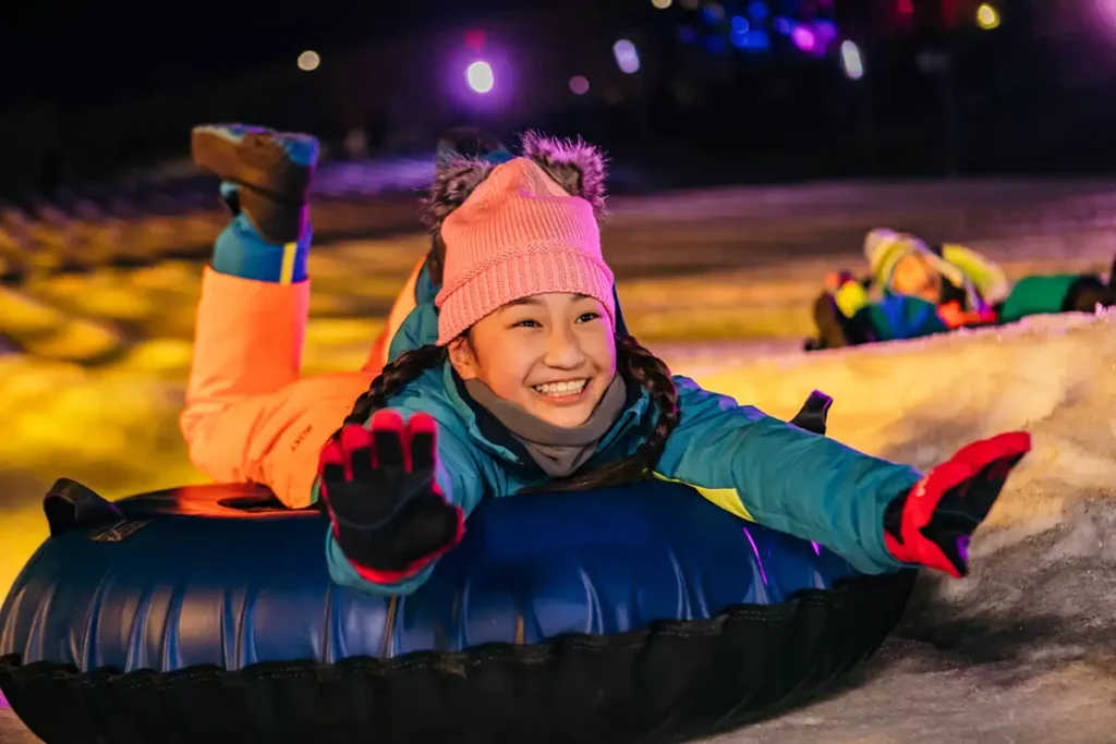 A young girl wearing a pink beanie with fuzzy ears, a colorful winter jacket, and gloves smiles excitedly as she slides down a snowy hill on a blue inflatable tube at night, with vibrant lights illuminating the scene and other riders in the background.